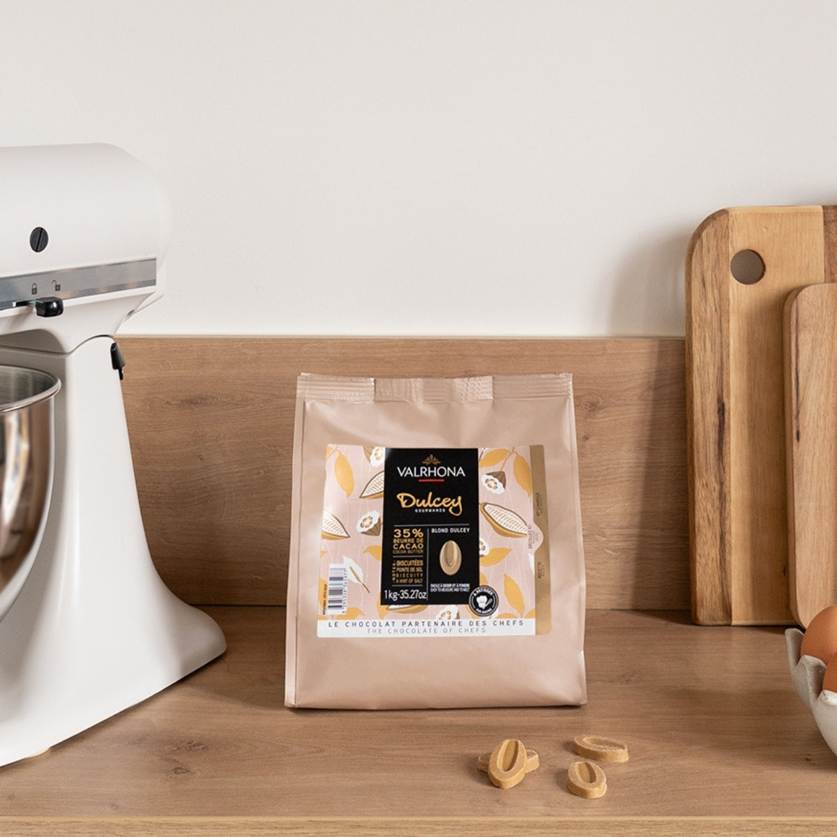 Photo ambiance sac de 1kg de chocolat Dulcey sur un plan de travail avec des fèves de chocolat, des oeufs et un robot de pâtisserie